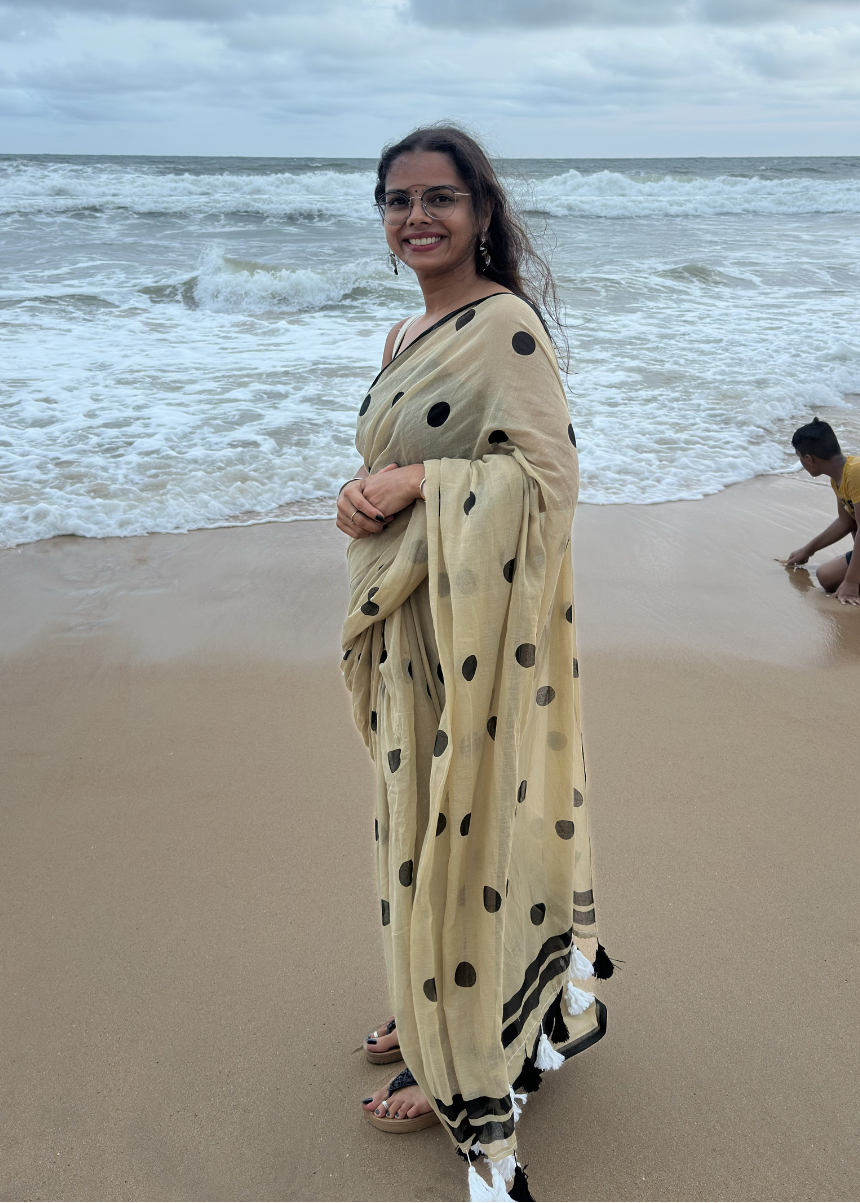 Woman in a beige saree with black polka dots standing on a beach with people playing in the water.